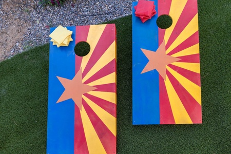 Two cornhole boards with a vibrant design featuring yellow rays, a red star in the center, and blue vertical stripes, placed on a green artificial turf. Two bean bags are on one of the boards.