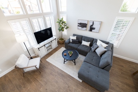 A modern living room with a grey sectional sofa, a round black coffee table, a TV on a white stand, a potted plant, wall art, and large windows with white blinds.