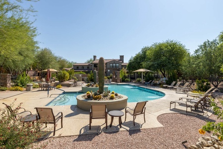 Outdoor swimming pool area with lounge chairs, umbrellas, a central cactus planter, and a building in the background surrounded by trees and desert landscaping.