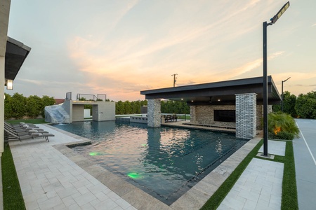 Outdoor modern pool area with adjacent covered patio and lounge chairs. The pool features a built-in waterfall and green lighting within the water. The sky is cloudy with hints of a sunset.