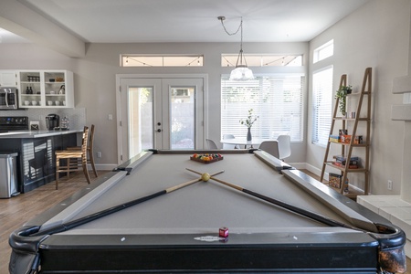 A room with a pool table in the center, kitchen area visible on the left, and a dining table by the window on the right. The walls have shelves with books and board games.