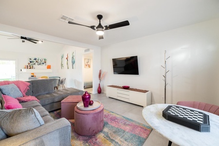 A modern living room with a gray sectional sofa, pink ottomans, colorful rug, wall-mounted TV, and ceiling fan. A small dining area and kitchen are visible in the background.