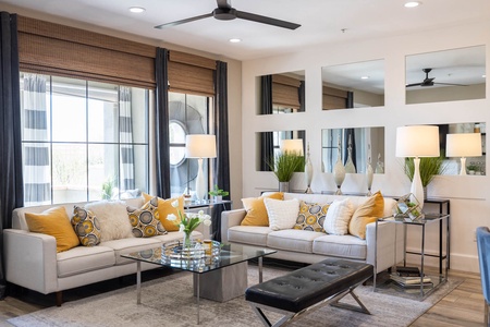 Modern living room with two beige sofas, yellow and patterned cushions, glass coffee table, black bench, tall lamps, decorative plants, and large mirrors on the wall.