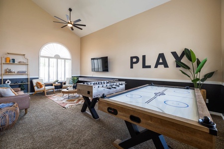 A game room with a foosball table, an air hockey table, a TV on the wall with the word "PLAY" above it, two chairs, a sofa, a small table, and multiple plants.