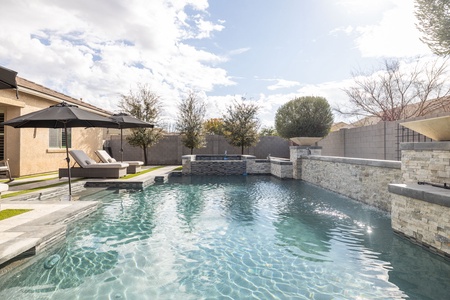 A backyard pool with a stone surrounding wall, lounge chairs under black umbrellas, and a small waterfall feature under a partly cloudy sky.