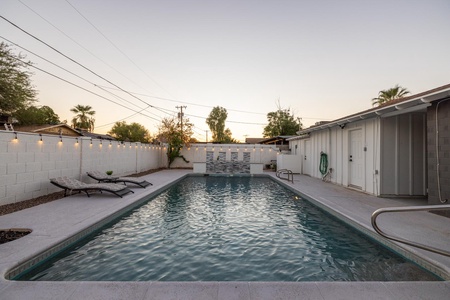 A backyard with a swimming pool, two lounge chairs, a white wall with light string, and a small waterfall feature under a clear sky at dusk.