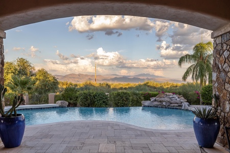 Covered backyard patio with a stone floor overlooking a pool and garden, with a scenic view of distant mountains and clouds in the background.