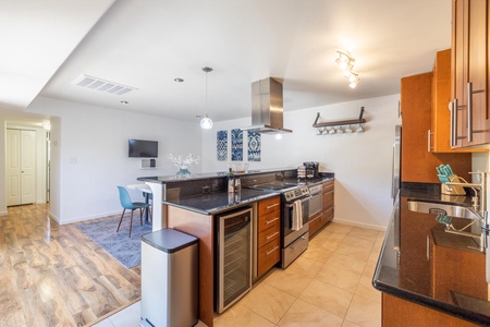 Modern kitchen with granite countertops, stainless steel appliances, wooden cabinets, and a dining area with a small table and blue chairs. A hallway and a bright room with wooden floor are visible.