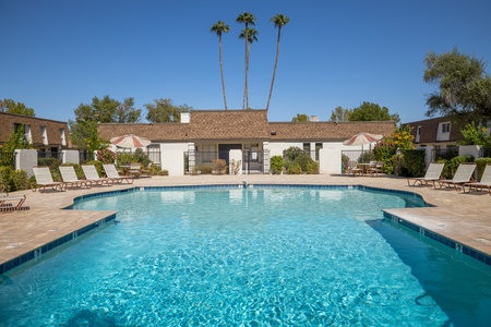 Outdoor swimming pool area with lounge chairs, umbrellas, and a central building in the background. The pool is surrounded by greenery and tall palm trees under a clear blue sky.