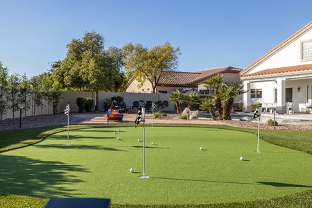 A backyard putting green with multiple holes and golf balls, surrounded by landscaping and a white house with patio furniture and trees in the background under a clear blue sky.