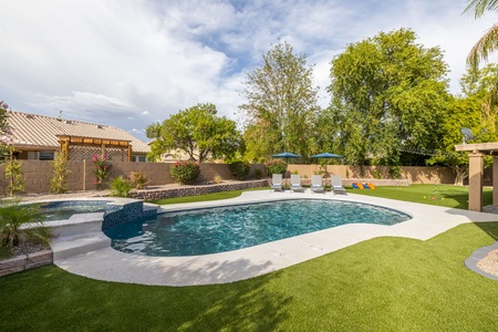 A backyard featuring a swimming pool with a waterfall, surrounded by patio lounge chairs, green lawn, trees, and a wooden pergola structure.