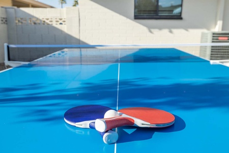 Two table tennis paddles, one red and one blue, lie side by side with two white balls on a blue ping pong table outside near a building.