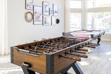 A game room with a foosball table in the foreground and a pool table in the background. The walls are adorned with framed photos and a coiled rope.