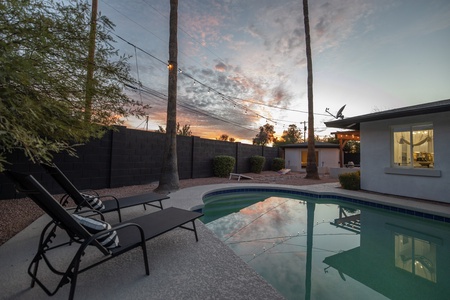 Poolside chairs and a tree-lined fence are silhouetted against a sunset sky, with a house and satellite dish visible in the background.