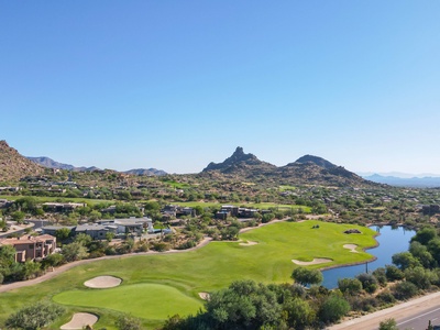 Aerial view of a golf course with a pond, surrounded by residential buildings and arid landscape, featuring rocky hills in the background under a clear blue sky.