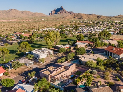 Aerial view of a residential area with sparse houses, green lawns, and desert landscape in the background, with a prominent rocky mountain in the distance under a clear blue sky.