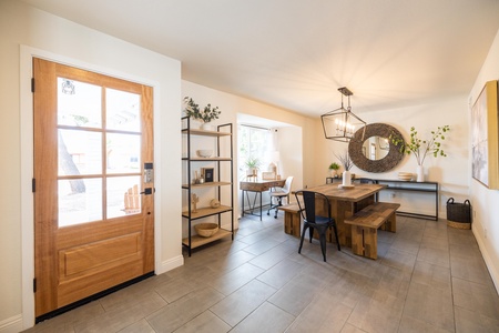 A well-lit dining area featuring a wooden table with black chairs, a desk in the corner with a chair, a large mirror on the wall, a cabinet with decorations, and a wooden entrance door.