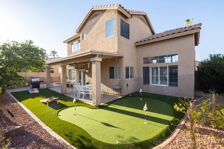 A two-story house with a beige exterior features an outdoor patio, dining area, and a small artificial grass putting green in a landscaped backyard.