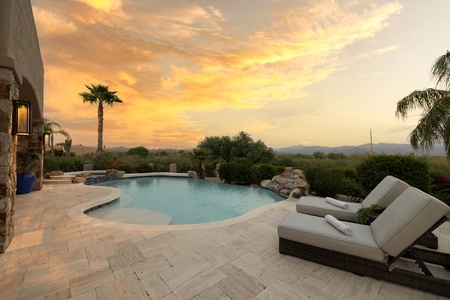 A serene outdoor pool area at sunset, featuring lounge chairs with towels, a palm tree, and lush greenery against a backdrop of mountains and a colorful sky.