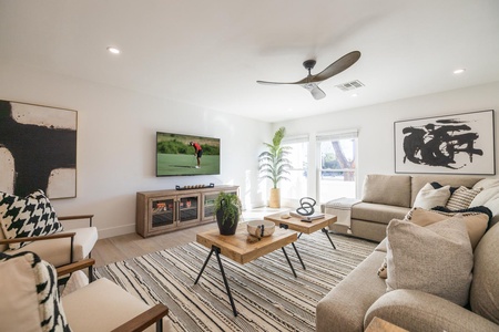 Modern living room with a large beige sectional sofa, wooden coffee table, wall-mounted TV, and abstract art. The room is well-lit with natural light from windows and features neutral decor and a ceiling fan.