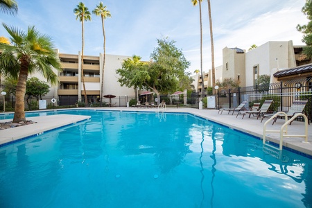 Outdoor swimming pool surrounded by lounge chairs and palm trees, with multi-story residential buildings in the background.