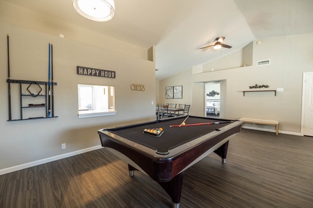 A well-lit recreation room with a black pool table, cue sticks on the wall, and a "Happy Hour" sign above a small window opening. There's also a dining area and a fan on the ceiling.