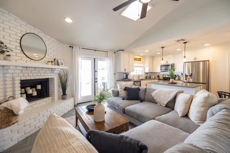 Modern living room and kitchen with white brick fireplace, gray sectional sofa, wood coffee table, stainless steel appliances, and recessed lighting. Natural light filters through glass double doors.