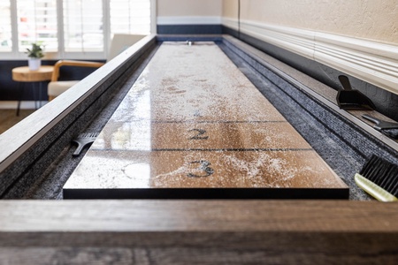 A close-up of a shuffleboard table with powdered surface and pucks, located indoors near a chair and a small table with a potted plant.