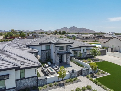 Aerial view of a modern, single-story house with a spacious patio, well-manicured lawn, and mountainous landscape in the background. Multiple similar houses surround the property.