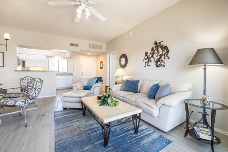 A living room with a white couch, blue pillows, and a coffee table on a blue rug. There is a ceiling fan, lamp, and wall art. The kitchen area is visible in the background.