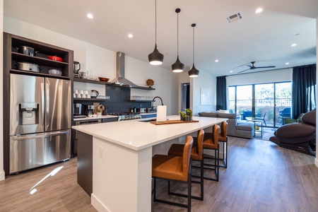 Modern kitchen and living area with stainless steel appliances, a large island with three brown chairs, pendant lights, a gray sectional sofa, ceiling fan, and sliding glass doors leading outside.