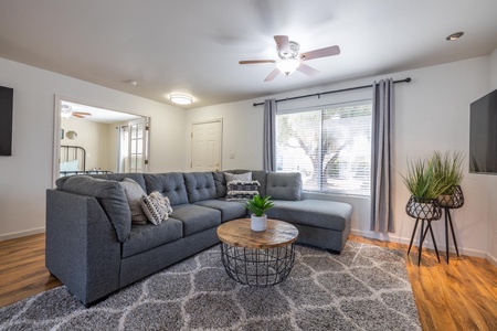 A modern living room with a gray sectional sofa, wooden coffee table, and two plants. The room features a ceiling fan, gray curtains and a large window letting in natural light.