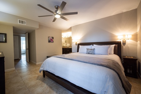 A modern bedroom with a large bed, two nightstands, and wall-mounted lamps. A ceiling fan hangs above and a doorway leads to an adjacent room. Neutral-colored decor with tiled floor.