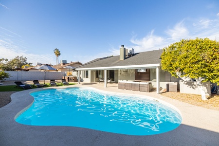 Backyard view of a house featuring a swimming pool, lounge chairs, a covered patio, and outdoor furniture, with a clear blue sky and palm trees in the background.