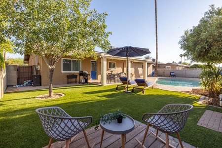 A backyard with a swimming pool, patio chairs, a table, a large umbrella, and a well-maintained lawn. A tree provides shade, and a house with a covered porch is in the background.