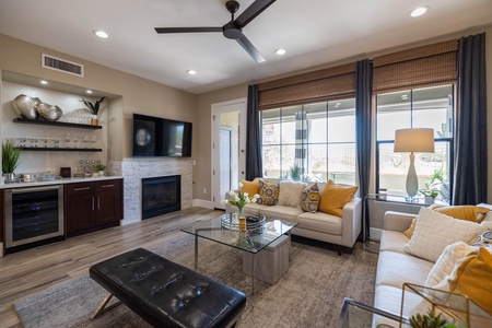 A modern living room with a ceiling fan, two sofas, a glass coffee table, and a wall-mounted TV above a fireplace. Large windows with drapes provide natural light; a bar area is on the left side.