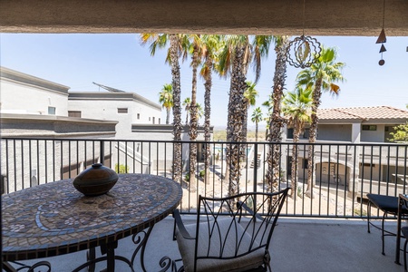 A covered balcony featuring a mosaic table with chairs overlooks a courtyard with palm trees and neighboring buildings.