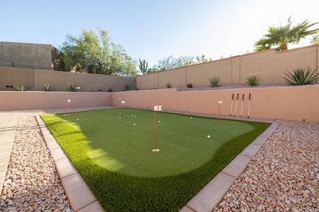 A backyard putting green is surrounded by desert landscaping with gravel and cacti. Golf clubs are placed against the wall, and several golf balls and flags are on the green.