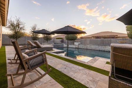 Outdoor pool area with rockers and loungers, shaded by umbrellas. Stone deck surrounds the pool, bordered by greenery and a stone wall. Sunset sky in the background.