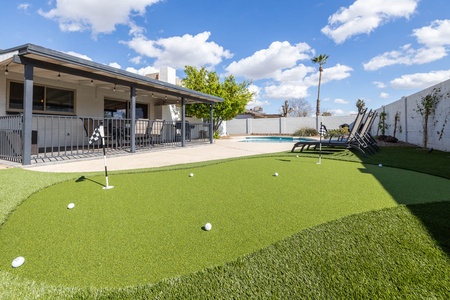 Backyard space with covered patio, miniature golf putting green, lounge chairs, trees, and a swimming pool.