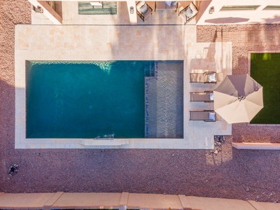 Aerial view of a rectangular swimming pool with clear water, adjacent lounge chairs, an umbrella, and a light-colored stone patio.