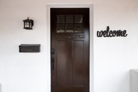 A dark brown wooden door flanked by a mailbox on the left and a lantern above it, with a "welcome" sign on the wall to the right.