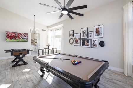 A modern game room features a pool table, foosball table, and a wall-mounted TV showing a football game. The walls display framed photos and a few cowboy hats.