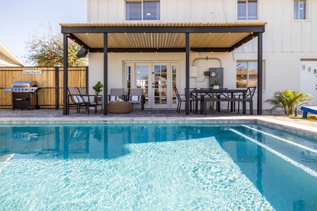 A backyard features a covered patio with seating, dining table, and grill, adjacent to a swimming pool with clear blue water.