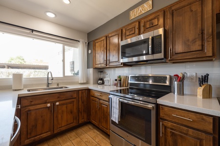 A kitchen with wooden cabinets, stainless steel appliances including a microwave and oven, a sink with a window above it, and various kitchen utensils on the countertops.