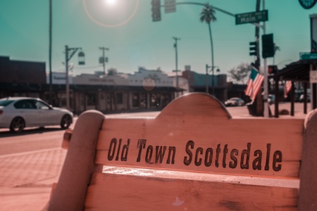 View of a wooden bench engraved with "Old Town Scottsdale" in a street scene with a car, buildings, traffic lights, and a palm tree in the background.