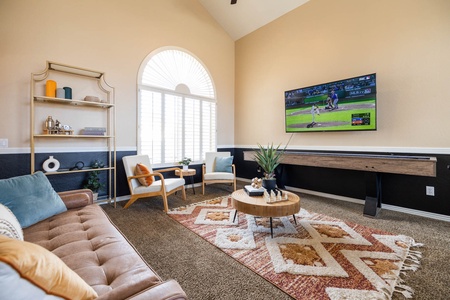 A living room with wooden shelves, a mounted TV showing a baseball game, a brown sofa, two white chairs, a round wooden coffee table, and a patterned rug on the carpeted floor.