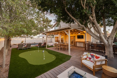 A backyard with a covered patio, outdoor furniture, bar area, putting green, trees, and a fire pit table.