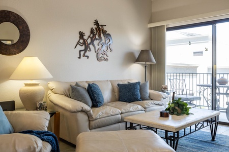 A well-lit living room with beige sofas, blue cushions, a coffee table, a mirror, decorative wall art, and a floor lamp. Sliding glass doors open to a balcony with outdoor furniture.