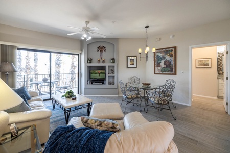 Living room with beige furniture, a coffee table, and a TV on a built-in shelf. A dining table with four chairs and a hanging lamp are on the right. Balcony view through sliding glass doors on the left.